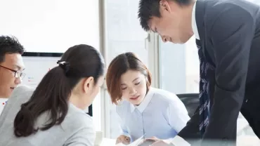 Group of people engaged in discussion at an office desk with a computer screen in the background.